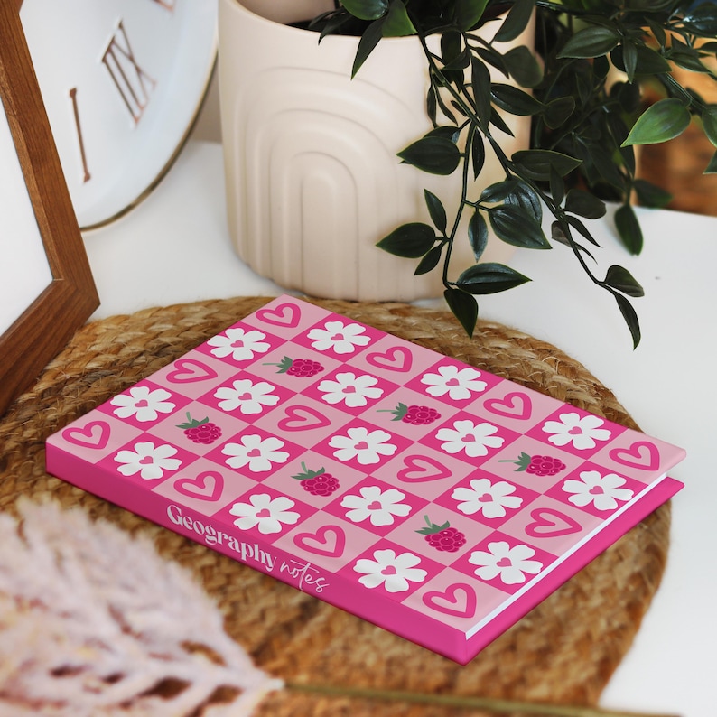 A raspberry and heart patterned hardcover notebook lying flat with the spine customised to say "Geography notes". The setting includes a woven mat, potted plant, and cozy neutral décor.