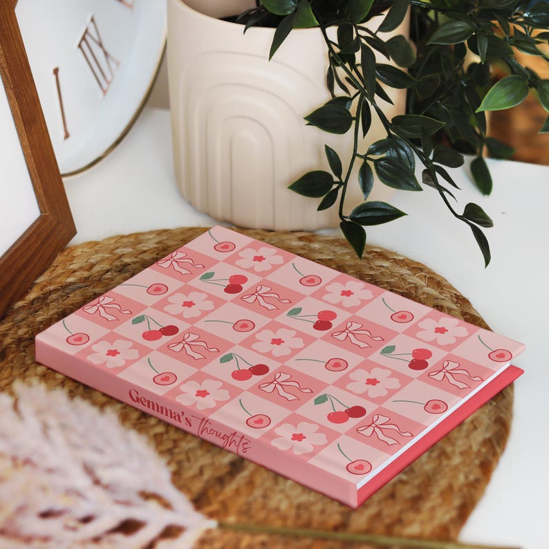 A flat-lay of a cherry-patterned hardcover notebook resting on a woven mat. The pink spine is personalised with the text "Gemma’s thoughts" in red cursive. Decorative plants and a clock are in the background.