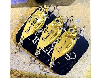 May include: Three gold-colored fishing lures with treble hooks, each with a different name in black script: "Bam Bam", "Pinchy", and "Dieter". The lures are on a black surface with white text.