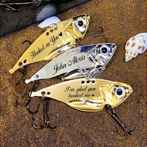 Könnte beinhalten: Nahaufnahme von drei personalisierten Angelködern. Zwei sind goldfarben und einer silberfarben. Die goldenen Köder tragen die Aufschriften "Hooked on You" und "I'm glad you hooked me". Der silberne Köder ist mit dem Namen "John Alexis" graviert.