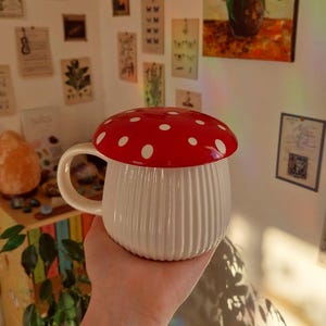 May include: A whimsical mushroom-shaped mug with a red lid dotted with white spots. The mug's body is white with vertical ribbed detailing. It has a curved handle and is designed to resemble a mushroom. A fun and unique kitchenware item.