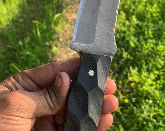 May include: A handcrafted knife with a textured, dark handle and a serrated edge. The blade is silver and angled, with a flat top. Two small rivets secure the handle. A leather lanyard is attached to the base of the handle.