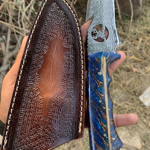 May include: A handcrafted knife with a patterned steel blade and a blue and gold handle. The knife is held next to a brown leather sheath with a leaf design. The blade has a decorative cutout.