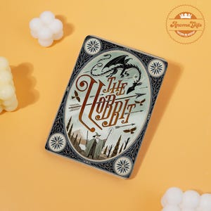 Könnte beinhalten: Ein rechteckiger Dekorationsartikel mit dem Titel "The Hobbit" in einer stilisierten Schriftart. Das Design umfasst Illustrationen eines Drachen, Pfeile und einen Zauberer. Der Rand ist dunkel mit floralen Akzenten, vor einem hellen Hintergrund.