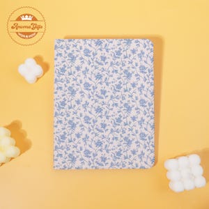 May include: A floral patterned notebook with a light blue and white design. The notebook has a rounded edge and is surrounded by white wax melts. The logo "Aroma Gifts" is in the upper left corner.