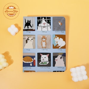 May include: A light blue notebook featuring a grid of nine panels, each displaying a different cat meme illustration. The illustrations include various cat poses and expressions, with text below each image. The notebook has a spiral binding.