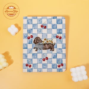 May include: A notebook with a blue and white checkered pattern and a cartoon dachshund wearing a striped shirt and carrying flowers. Red cherries are scattered across the notebook's surface. The notebook has rounded corners.