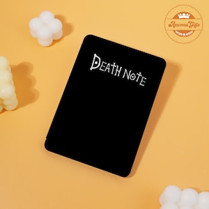 May include: A black rectangular notebook with the words "Death Note" in white script. The notebook is on a light orange surface, with white bubble-shaped candles in the background. The notebook is a product from Aroma Gifts.