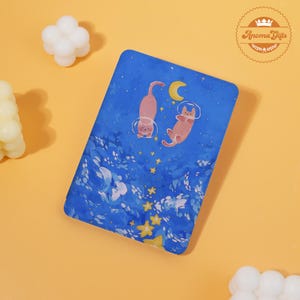 May include: A blue rectangular item with a whimsical illustration of two pink cats in space suits floating among stars and a crescent moon. The item has a textured surface and is set against a yellow background. The item is branded with the words "Aroma Gifts".