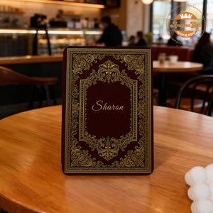 May include: A rectangular dark maroon electronic reader cover featuring an intricate gold filigree border and the name Sharon printed in a white script font in the center. The cover has a textured surface and a classic, ornate decorative design.