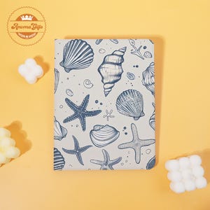 May include: A notebook with a light gray cover featuring a pattern of blue seashells and starfish. The notebook has a cream-colored background and the text "Aroma Gifts" in a circular logo.
