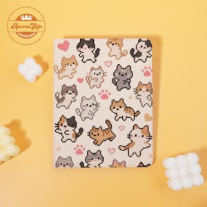 May include: A notebook with a white cover featuring a repeating pattern of pixelated cats in various colors, pink hearts, and paw prints. The notebook has rounded corners and is set against a yellow background. The brand logo "Aroma Gifts" is in the upper left corner.