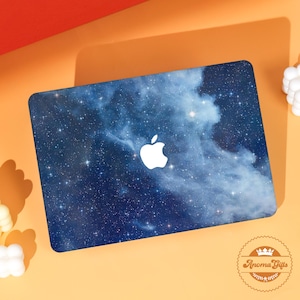 Könnte beinhalten: Ein Laptop mit blauem Galaxiemuster. Die Hülle zeigt einen Sternenhimmel mit weißen Wolken und einem weißen Apple-Logo. Der Laptop befindet sich auf einer orangefarbenen Oberfläche.