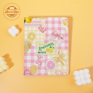 May include: A decorative notebook with a pink and white checkered pattern, featuring lemon and bow illustrations. The notebook has the text "Fresh Lemonade Sold Here" and "XOXO". The overall theme is a summer or spring aesthetic.