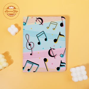 May include: A notebook with a musical theme. The cover features a pattern of black musical notes, treble clefs, and other symbols on a light blue and pink striped background. The notebook has a rounded corner design.