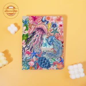 May include: A notebook with a colorful marine life design. The cover features illustrations of jellyfish, starfish, sea turtles, and flowers in shades of pink, blue, and green. The notebook has a light gray background.