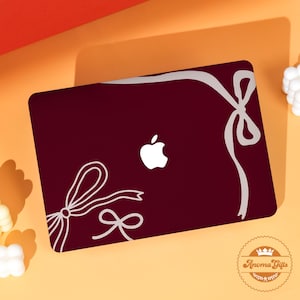 Puede incluir: Funda para portátil de color burdeos con el logotipo blanco de Apple y diseños de lazos de cinta blancos. La funda tiene un acabado brillante y está diseñada para un portátil. El logotipo de Anoma Gifts está en la esquina inferior derecha.