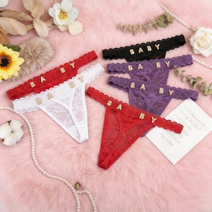 May include: A collection of lace thong underwear in red, white, black, and purple. Each thong features the word "BABY" spelled out in gold-colored letters across the front. The underwear is displayed on a pink, fluffy surface.