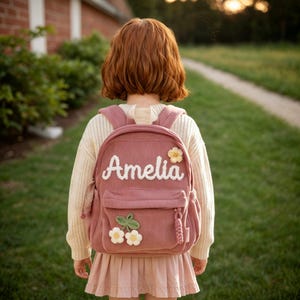 Puede incluir: Una mochila de pana color rosa empolvado con el nombre Amelia en letras blancas bordadas, una pequeña aplicación de flor amarilla y un tallo verde con dos flores blancas en el bolsillo delantero, sobre un suéter de punto color crema.