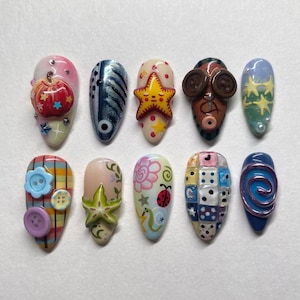 May include: Ten artificial nails, each with a distinct design. The collection includes nails with a red apple, a fish, a starfish, and a face with button eyes. Other designs feature flowers, buttons, and swirls, offering a variety of styles.