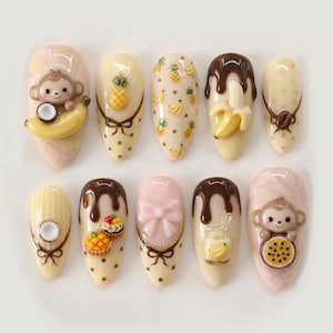 May include: A collection of ten almond-shaped artificial nails featuring 3D designs. The designs include a monkey with a banana, a pineapple, bananas, chocolate drips, and a pink bow. The nails are painted in pastel colours.