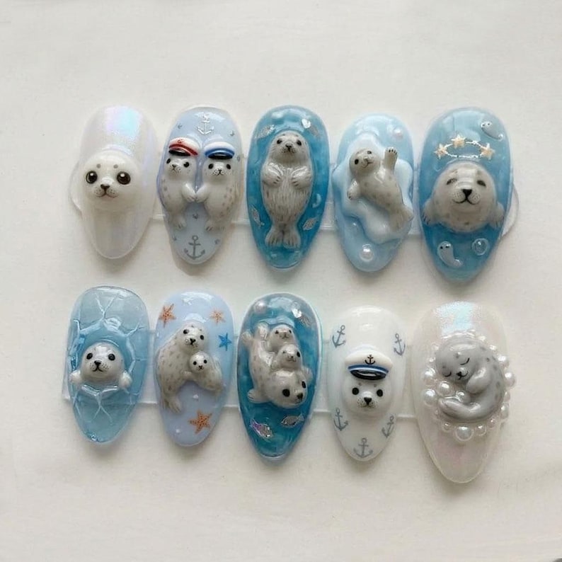 May include: A set of ten artificial nails with 3D seal designs. Each nail features a different seal design, including seals wearing sailor hats, swimming in water, and resting. The nails are painted in shades of blue, white, and iridescent colors.