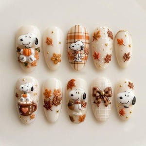 May include: A set of ten press-on nails featuring a cream base with 3D Snoopy figures, orange and brown autumn leaves, plaid patterns, a brown mug with a heart, and a brown checkered bow. The designs include gold accents and small dots.