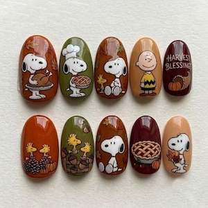 Op de afbeelding: Een set van tien kunstnagels met Peanuts-figuren in herfstthema. De ontwerpen bevatten Snoopy met een kalkoen, een taart en bladeren, Charlie Brown, Woodstock, eikels, pompoenen en de tekst Harvest Blessings in bruin, oranje en groen.