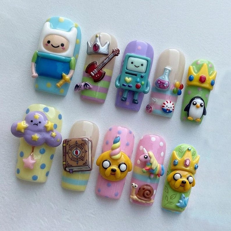 May include: A set of ten artificial nails with cartoon character designs. Each nail features a different character, including Finn, BMO, Jake the Dog, and a penguin. The nails are painted in various colors, such as blue, pink, and green, with polka dots and stripes.