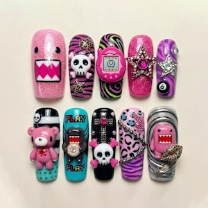 Könnte beinhalten: Eine Sammlung von zehn künstlichen Nägeln mit verschiedenen Designs. Die Nägel zeigen rosa Glitzer, Zebrastreifen, Totenköpfe, ein Tamagotchi, Sterne und einen rosa Bären. Einige Nägel haben das Wort "PLAY" und einen Domo-kun-Charakter.
