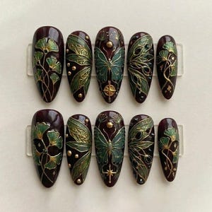 May include: A set of ten almond-shaped false nails. Each nail is a deep burgundy color with intricate designs in green and gold. The designs include butterflies, leaves, and floral patterns, with small gold accents.