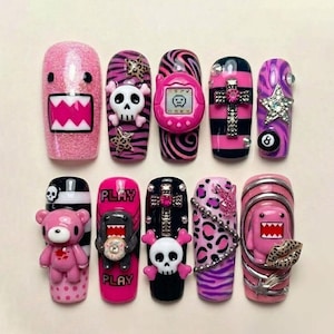 Könnte beinhalten: Eine Sammlung von Kunstnägeln mit verschiedenen Designs. Die Nägel zeigen rosa Glitzer, Cartoon-Charaktere, Totenköpfe, ein Tamagotchi, ein Kreuz, einen Stern und eine 8-Ball. Einige Nägel tragen das Wort "PLAY". Die Nägel sind in verschiedenen Formen und Größen.