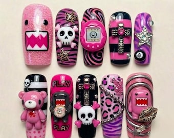 Op de afbeelding: Een verzameling kunstnagels met diverse ontwerpen. De nagels tonen roze glitter, stripfiguren, schedels, een Tamagotchi, een kruis, een ster en een 8-ball. Sommige nagels hebben het woord "PLAY". De nagels zijn in verschillende vormen en maten.