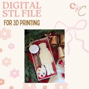 Puede incluir: Una caja transparente contiene una figura de cascanueces blanca y un objeto en forma de paleta con salpicaduras de pintura coloridas. La caja está atada con una cuerda dorada y una etiqueta que dice "Merry Christmas". El texto "DIGITAL STL FILE FOR 3D PRINTING" está en la parte superior.