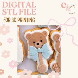 May include: A digital STL file for 3D printing featuring a teddy bear rattle cookie design. The cookie has a brown bear head with a light blue bow, set against a white and tan background. The text "DIGITAL STL FILE FOR 3D PRINTING" is at the top.