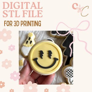 May include: Digital STL file for 3D printing. The image shows a close-up of a yellow smiley face cookie with black lightning bolt eyes and a curved smile. Other cookies are visible, including a party hat and a black and white checkered design.