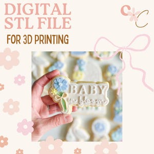 Könnte beinhalten: Eine digitale STL-Datei für den 3D-Druck. Das Bild zeigt einen dekorierten Keks mit den Worten "BABY in bloom" und einem floralen Design. Der Keks wird in einer Hand gehalten, mit anderen dekorierten Keksen im Hintergrund. Der Hintergrund ist hellbeige mit rosa floralen Akzenten.