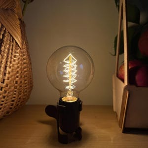 Könnte beinhalten: Eine klare, runde Glühbirne aus Glas mit einem sichtbaren, leuchtenden Spiralfaden im Inneren. Die Glühbirne ist in einer dunklen, mattschwarzen Metallsockelfassung montiert, die einen kleinen, hervorstehenden Hebel oder Schalter besitzt.
