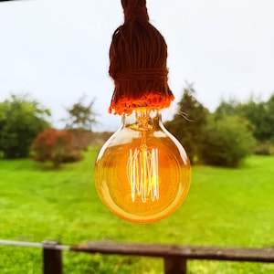 Könnte beinhalten: Eine runde, bernsteinfarbene Glühbirne aus Glas mit sichtbarer vertikaler Filamentstruktur. Der obere Teil der Birne ist von einer texturierten, dunkelbraunen Jute- oder Schnurquaste umhüllt, die den Sockel bedeckt und in ein Aufhängeseil übergeht.