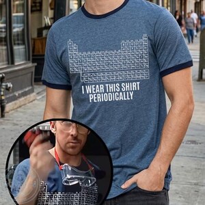 May include: A blue ringer t-shirt featuring a periodic table design and the text "I WEAR THIS SHIRT PERIODICALLY" in white. The shirt has a navy blue collar and sleeve trim. The shirt is made of a soft, comfortable material.