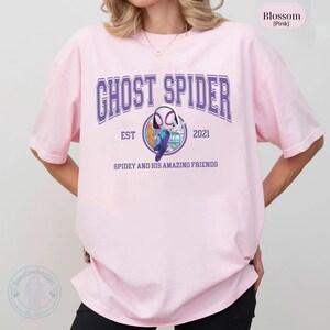 May include: A light pink t-shirt with the text "GHOST SPIDER" in purple block letters. Below the text is a graphic of a character from the Spider-Man universe. The shirt also has the text "EST 2021" and "SPIDEY AND HIS AMAZING FRIENDS".