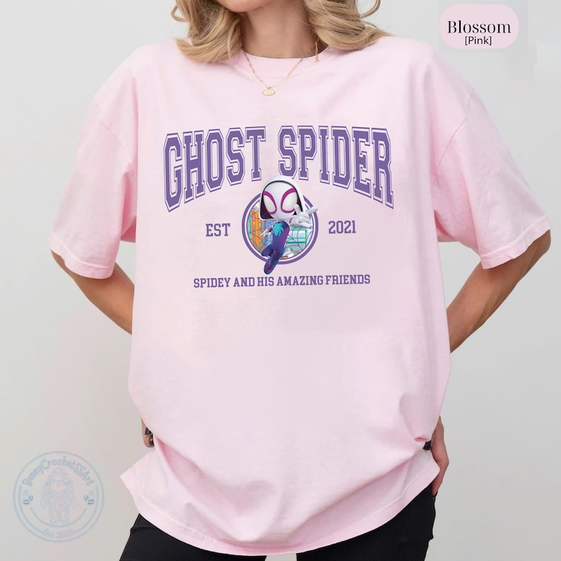 May include: A light pink t-shirt with the text "GHOST SPIDER" in purple block letters. Below the text is a graphic of a character from the Spider-Man universe. The shirt also has the text "EST 2021" and "SPIDEY AND HIS AMAZING FRIENDS".