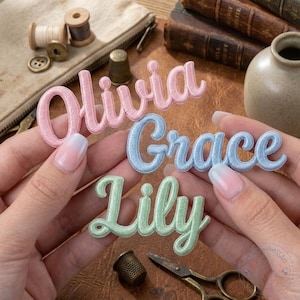 May include: Three embroidered name patches in pastel colours: pink, blue, and green. The names "Olivia", "Grace", and "Lily" are shown in a cursive script. Sewing accessories such as a thimble and scissors are also visible.