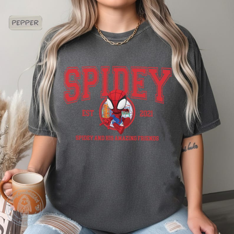 May include: Dark gray t-shirt featuring the word "SPIDEY" in red, with "EST 2021" below. A cartoon Spidey character is in the center. The text "SPIDEY AND HIS AMAZING FRIENDS" is below the character.