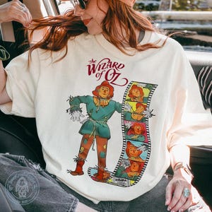 May include: Cream-coloured t-shirt featuring a graphic of the Scarecrow from The Wizard of Oz. The Scarecrow is depicted in a teal outfit with orange patches and a matching hat. The shirt also displays the film's title and a film strip.
