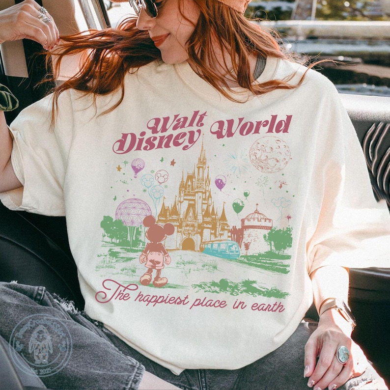 Puede incluir: Camiseta color crema con un gr&aacute;fico del castillo de Cenicienta, Mickey Mouse y el texto "Walt Disney World" y "The happiest place in earth" en rosa y verde. El dise&ntilde;o incluye globos, fuegos artificiales y un monorra&iacute;l.
