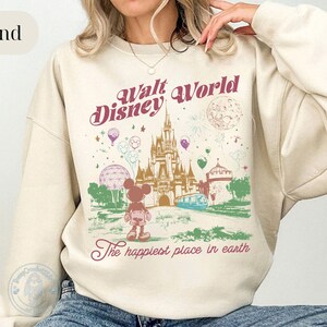 Puede incluir: Sudadera de color arena con un gr&aacute;fico de estilo vintage de Walt Disney World. El dise&ntilde;o incluye el castillo, Mickey Mouse, globos y el texto "Walt Disney World" y "The happiest place in earth".
