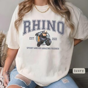 May include: Ivory-colored t-shirt featuring the word "RHINO" in a blue, collegiate-style font above an illustration of a rhino character. The text "EST 2021" and "SPIDEY AND HIS AMAZING FRIENDS" are also printed on the shirt.