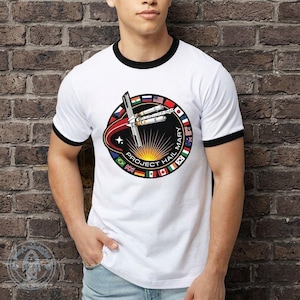May include: White ringer t-shirt with black trim featuring a circular graphic with a spaceship design, flags, and the text "PROJECT HAIL MARY". The design includes a star and a red and orange arc.