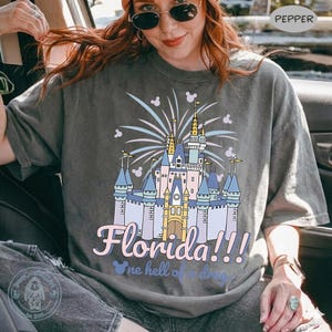 Puede incluir: Camiseta gris con una colorida ilustraci&oacute;n de un castillo, fuegos artificiales y la palabra "Florida!!!" en una fuente l&uacute;dica. El dise&ntilde;o incluye iconos de Mickey Mouse, creando un tema caprichoso.
